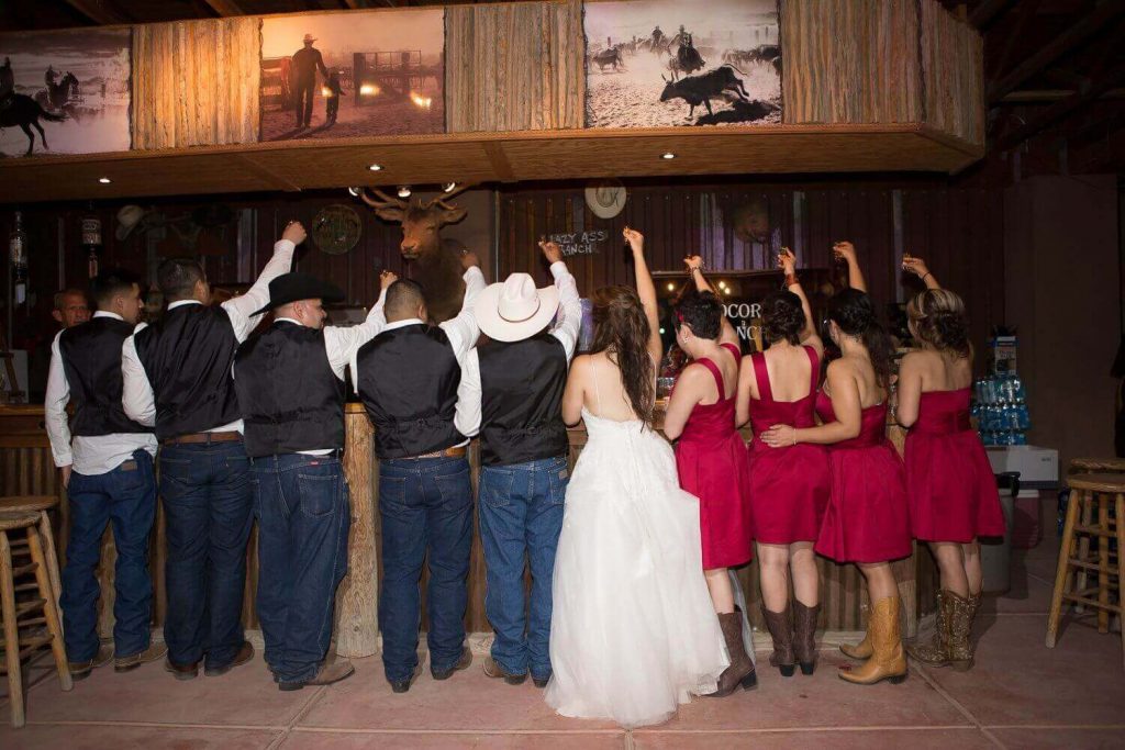 Wedding party lined up at the rustic bar raising shots—cowboy hats, red dresses, and western décor—inside the Pavilion at Cocoraque Ranch in Marana near Tucson.