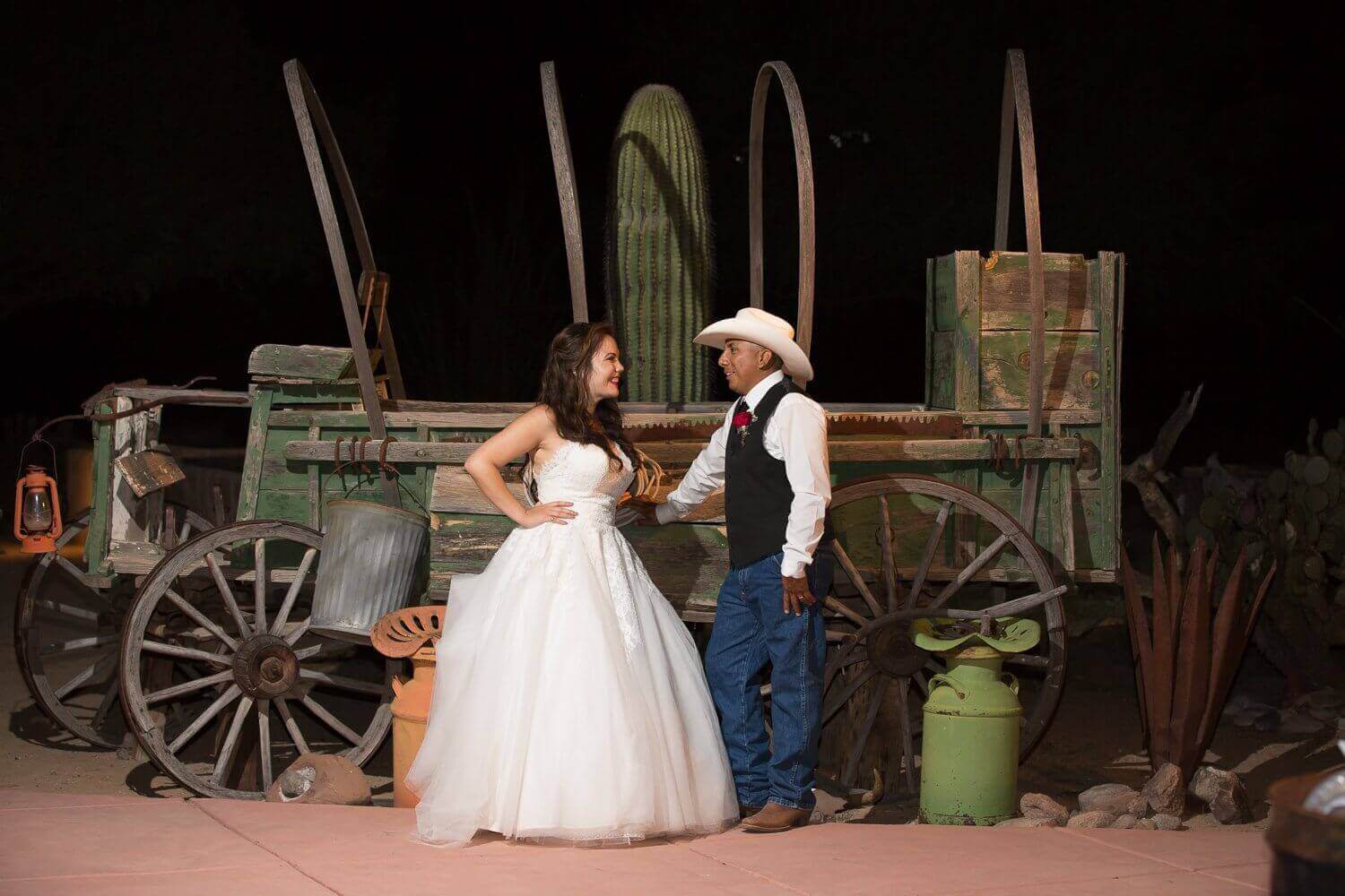 Bride in ball-gown and groom in cowboy hat posing by an antique green wagon with saguaro backdrop at Cocoraque Ranch in Marana near Tucson.