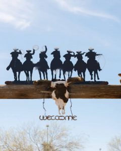 Silhouette of cowboys on horseback above a rustic wooden ranch entrance with a steer skull and a “Welcome” sign at Cocoraque Ranch.