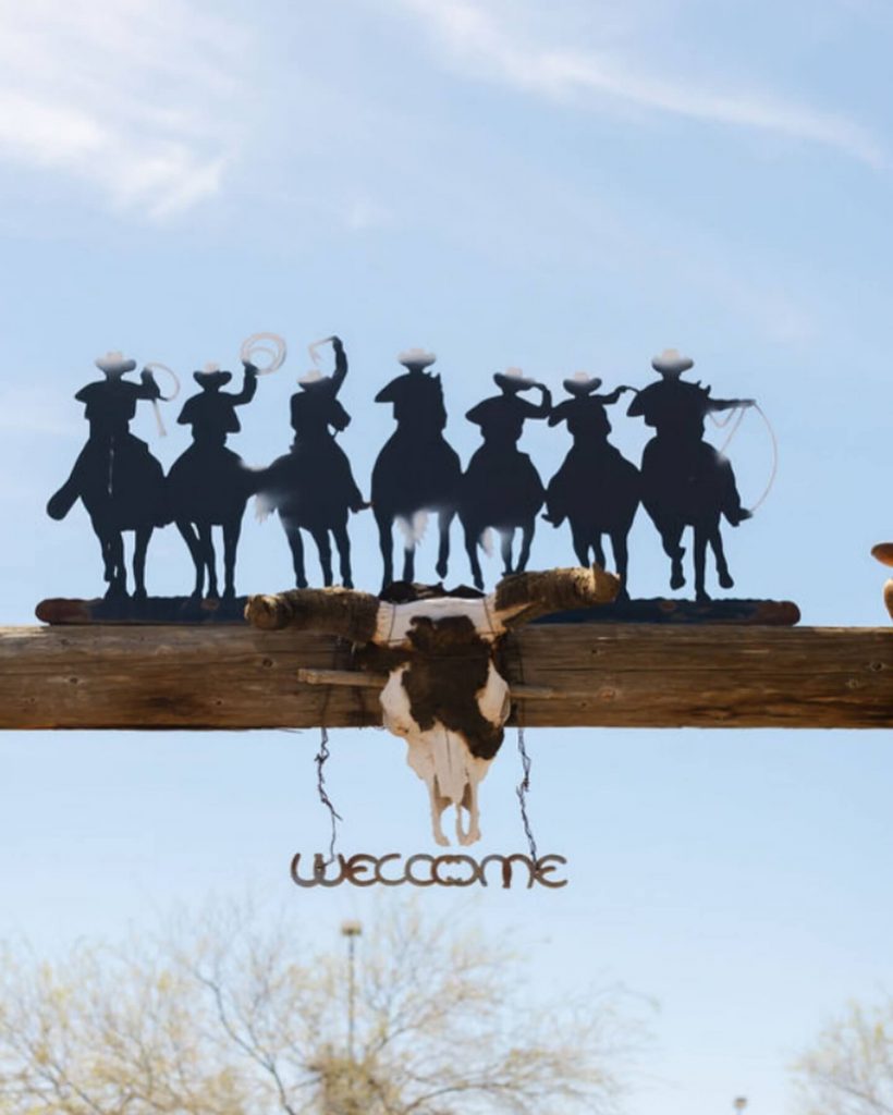 Silhouette of cowboys on horseback above a rustic wooden ranch entrance with a steer skull and a “Welcome” sign at Cocoraque Ranch.